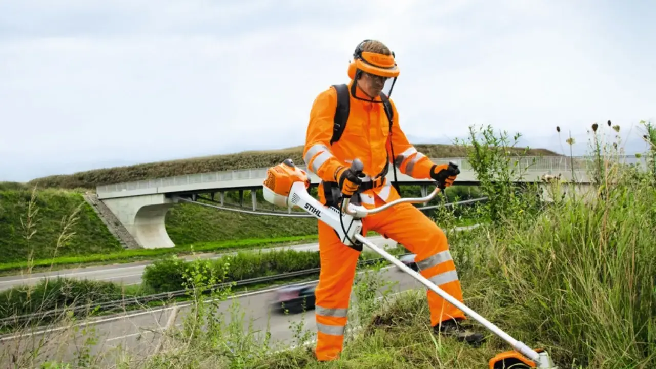 Pracownik w pomarańczowym stroju ochronnym z kosiarką spalinową Stihl, gotowy do pracy. Może to być pomocne przy pytaniu jak ustawić zapłon w kosiarce.