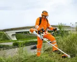 Pracownik w pomarańczowym stroju ochronnym z kosiarką spalinową Stihl, gotowy do pracy. Może to być pomocne przy pytaniu jak ustawić zapłon w kosiarce.