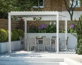 Nowoczesna pergola z jadalnianym zestawem w ogrodzie, otoczona kwitnącymi roślinami i zielenią. Idealne miejsce na relaks.