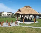 Altana z bali ze słomianym dachem, stołem i krzesłami w środku, otoczona zielenią i kamiennym kręgiem z pni.