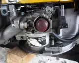 Zbliżenie na gaźnik kosiarki spalinowej z czerwonym przyciskiem "Cold Start PRIMER". Widoczne elementy mechanizmu, które mogą być pomocne, gdy zastanawiasz się, jak ustawić obroty w kosiarce spalinowej.