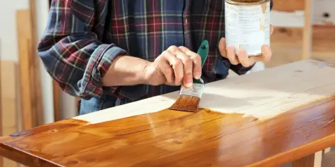 Osoba w kratę maluje pędzlem drewnianą ławkę lakierobejcą.
