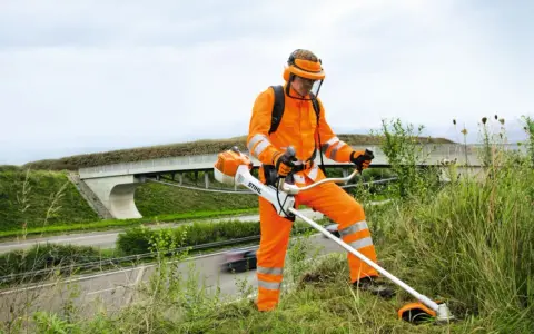 Pracownik w pomarańczowym stroju ochronnym z kosiarką spalinową Stihl, gotowy do pracy. Może to być pomocne przy pytaniu jak ustawić zapłon w kosiarce.