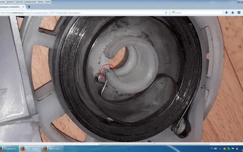 Szczegółowy widok mechanizmu szarpaka kosiarki, pokazujący jak założyć sprężynę do szarpaka kosiarki. Czerwony okrąg zaznacza punkt zaczepienia.