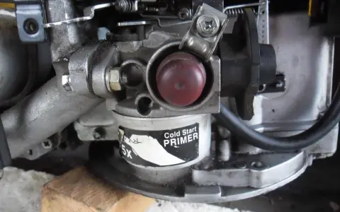 Zbliżenie na gaźnik kosiarki spalinowej z czerwonym przyciskiem "Cold Start PRIMER". Widoczne elementy mechanizmu, które mogą być pomocne, gdy zastanawiasz się, jak ustawić obroty w kosiarce spalinowej.