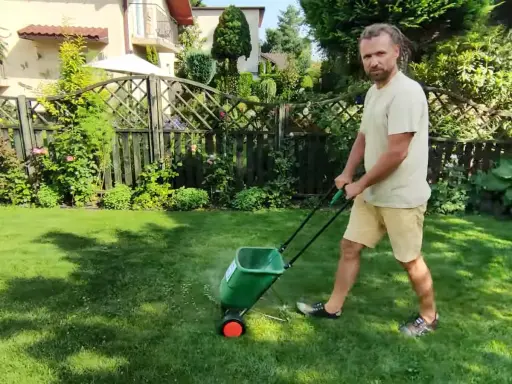 Mężczyzna z dredami używa zielonego siewnika do nawozu na trawniku. Idealny siewnik do nawozu na trawnik ułatwia pielęgnację ogrodu.