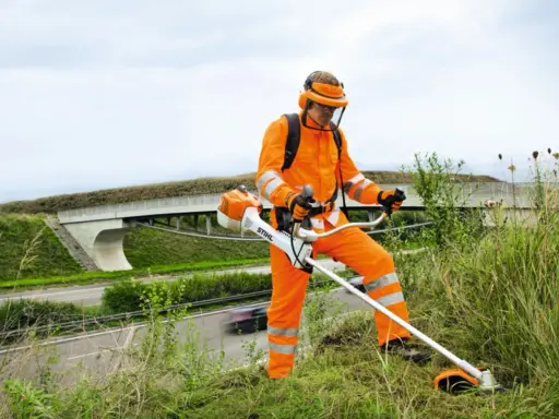 Pracownik w pomarańczowym stroju ochronnym z kosiarką spalinową Stihl, gotowy do pracy. Może to być pomocne przy pytaniu jak ustawić zapłon w kosiarce.