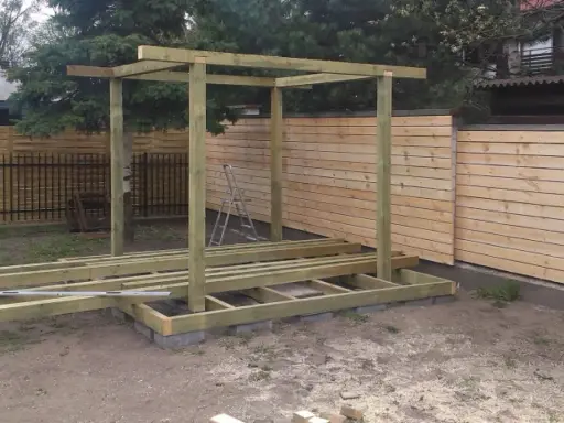 Konstrukcja altany w trakcie budowy. Drewniany szkielet i podłoga na bloczkach. W tle płot.