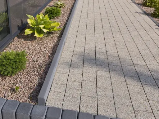 Nowoczesne obrzeża w ogrodzie wykonane z szarego betonu, oddzielające ścieżkę z kostki od rabaty z kamykami i roślinami.