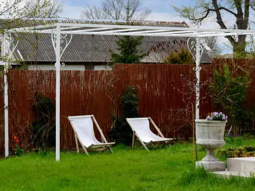 Biała pergola ogrodowa z dwoma leżakami i donicą z kwiatami.