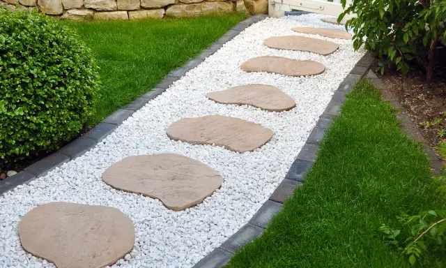 Ścieżka z białych kamieni i płyt, jak wysypać kamienie w ogrodzie, prowadzi do białej furtki. Obok trawnik i kamienny murek.