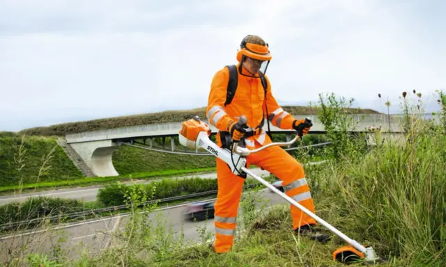 Pracownik w pomarańczowym stroju ochronnym z kosiarką spalinową Stihl, gotowy do pracy. Może to być pomocne przy pytaniu jak ustawić zapłon w kosiarce.