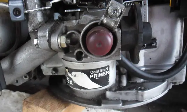 Zbliżenie na gaźnik kosiarki spalinowej z czerwonym przyciskiem "Cold Start PRIMER". Widoczne elementy mechanizmu, które mogą być pomocne, gdy zastanawiasz się, jak ustawić obroty w kosiarce spalinowej.