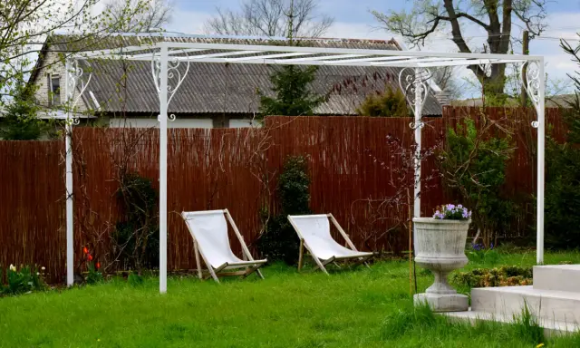 Biała pergola ogrodowa z dwoma leżakami i donicą z kwiatami.