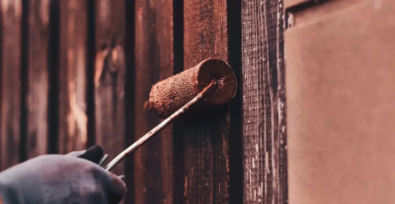 Ręka w rękawicy maluje drewnianą altanę brązową farbą. Idealny spos&oacute;b, czym pomalować altanę drewnianą, by ją odnowić.